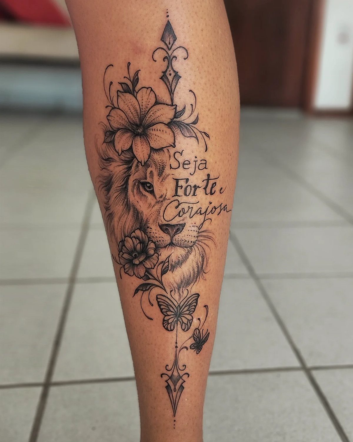 Black line tattoo on a leg featuring a lion, flowers, and cursive text.