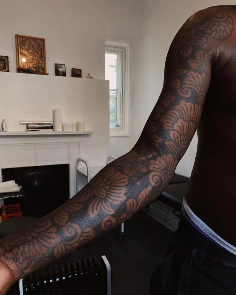 Brown-skinned arm with a detailed dark ink floral and wave pattern tattoo.