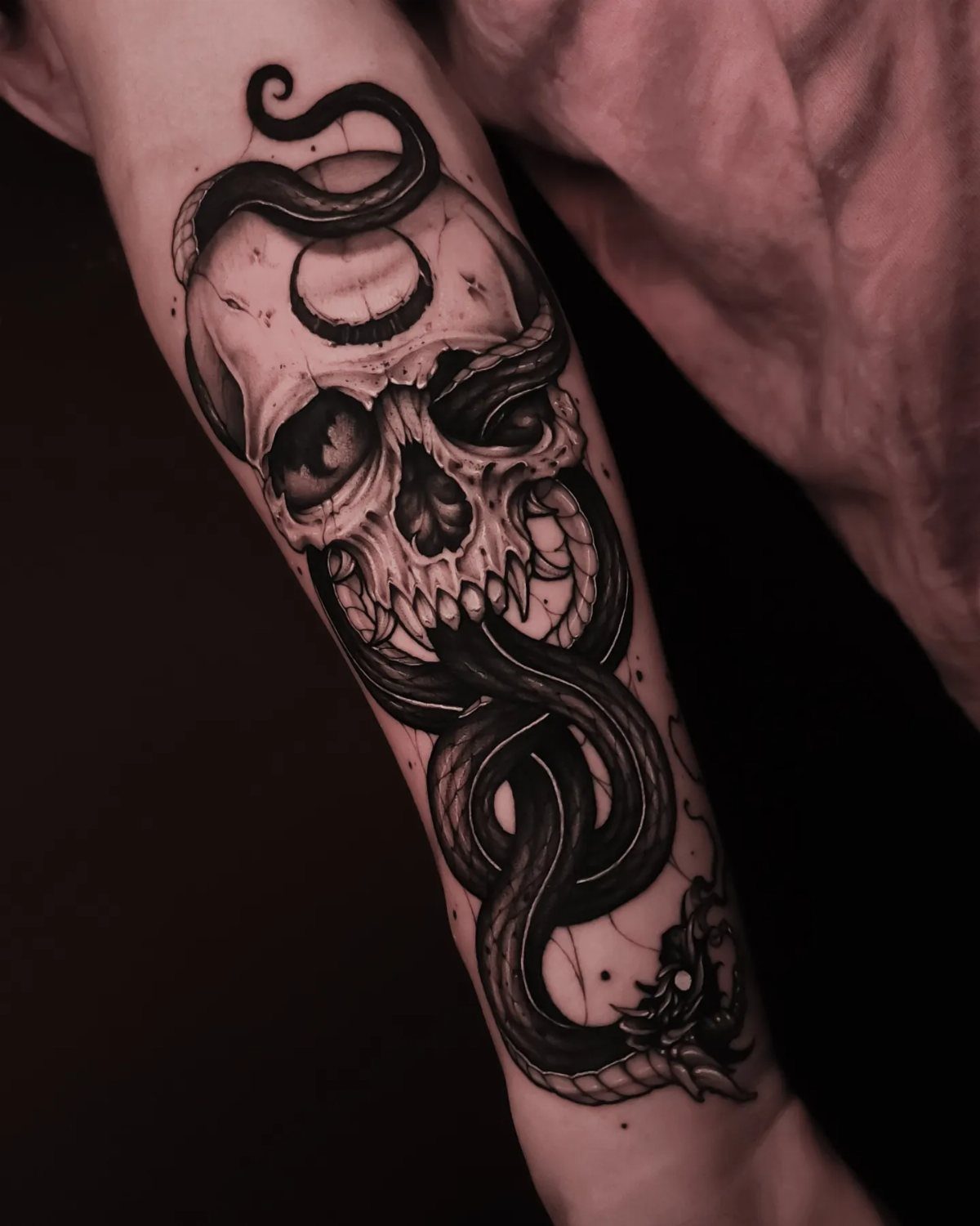 Skull and snake tattoo on a forearm in black and grey ink.