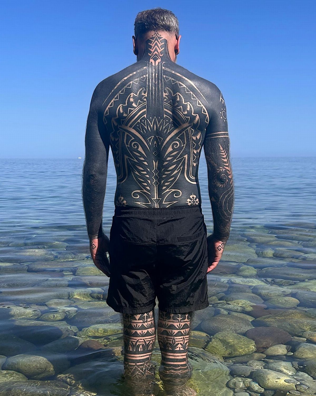 Man with large black ink tattoos on back and legs standing in shallow water