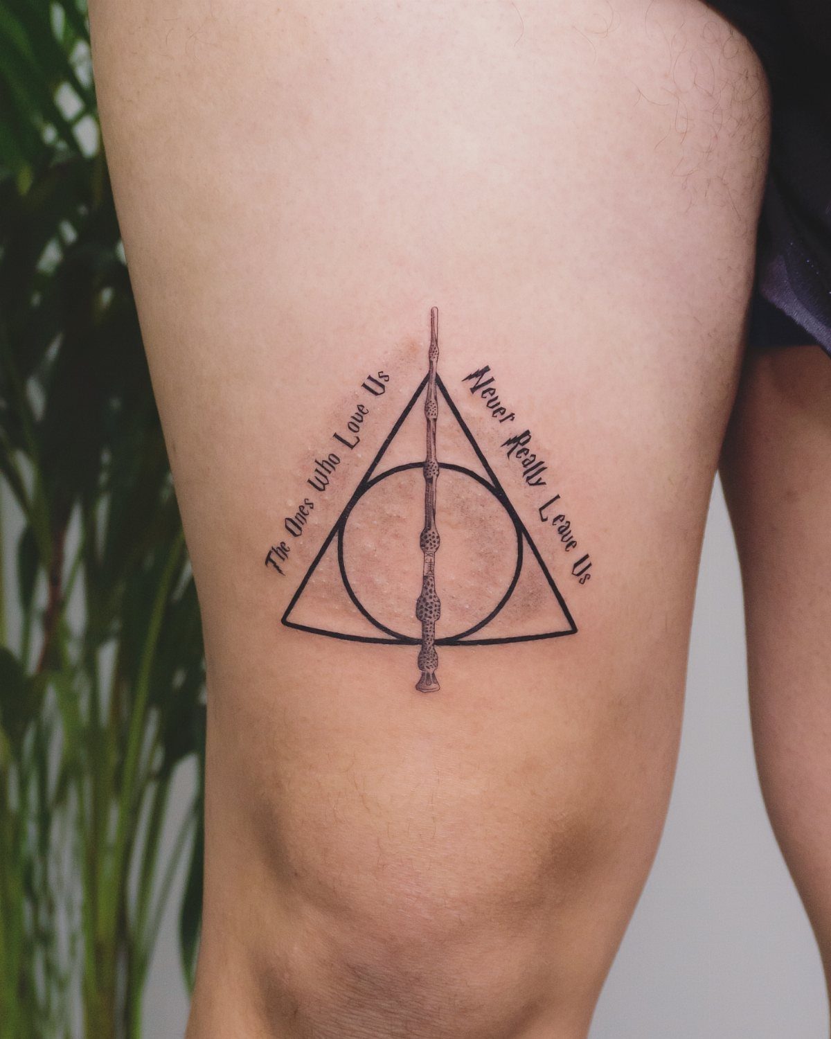 Deathly Hallows tattoo on a thigh with a sentimental quote in black ink.