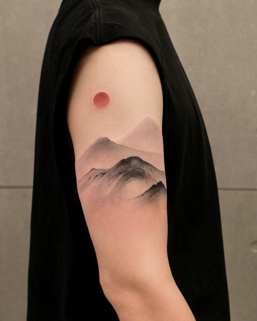 Tattoo on an arm showing a red rising sun over misty grey mountains.