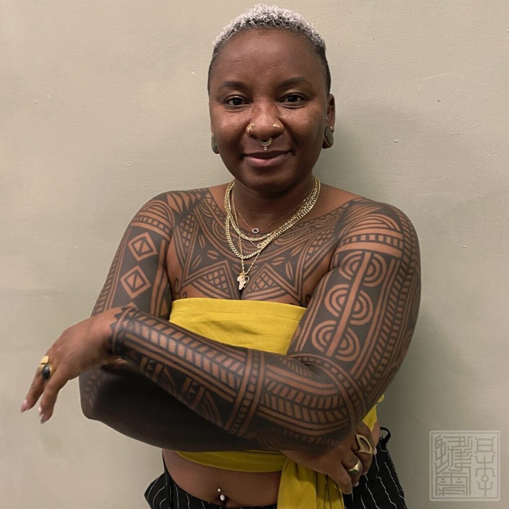 Woman with gold jewelry and yellow top showing intricate black geometric arm tattoos.