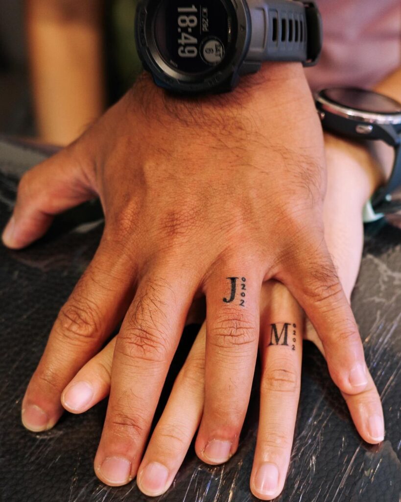 Two hands with small black ink wedding ring tattoos on their ring fingers.
